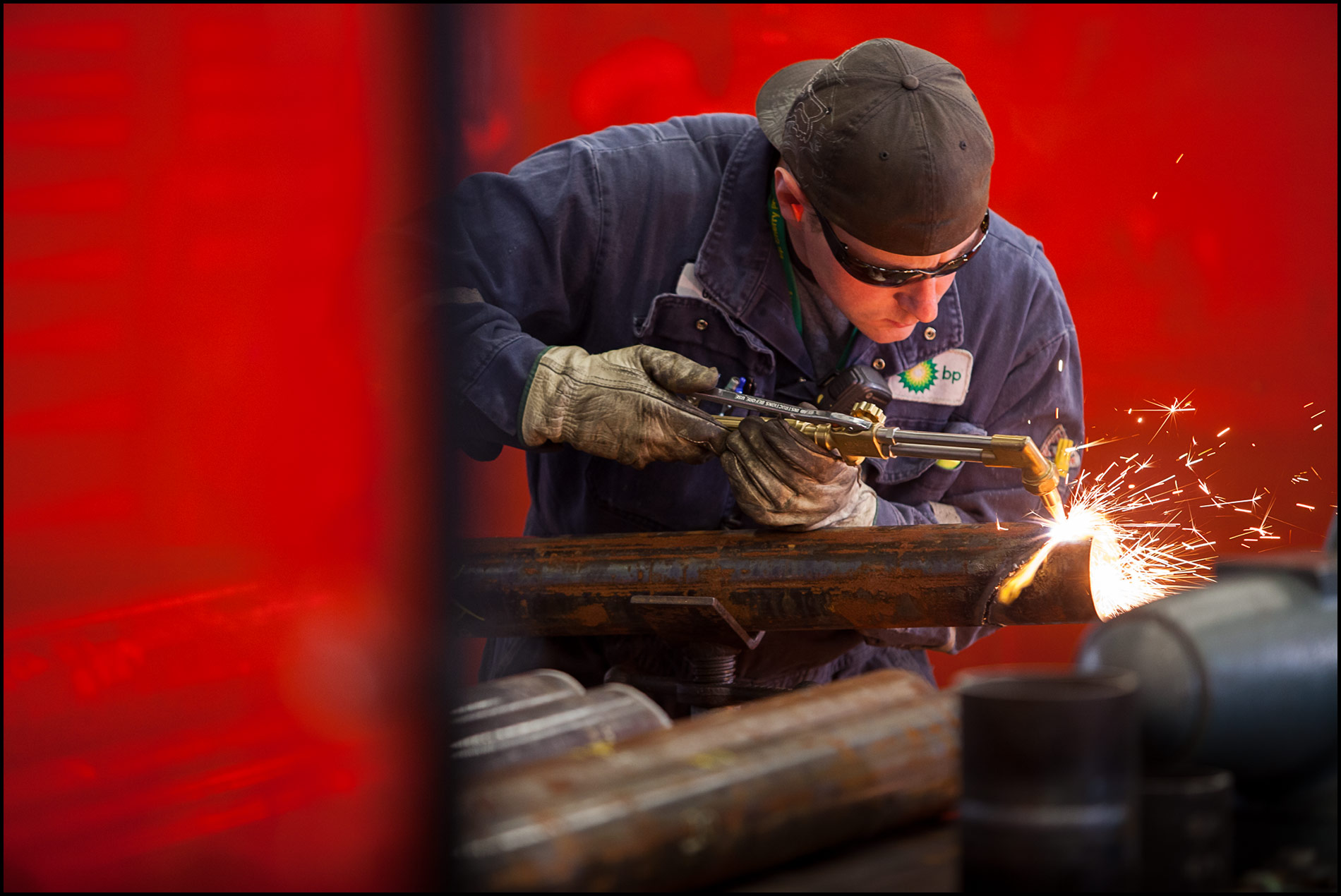 A welder uses a cutting torch behind red safety barriers in a refinery fabrication shop