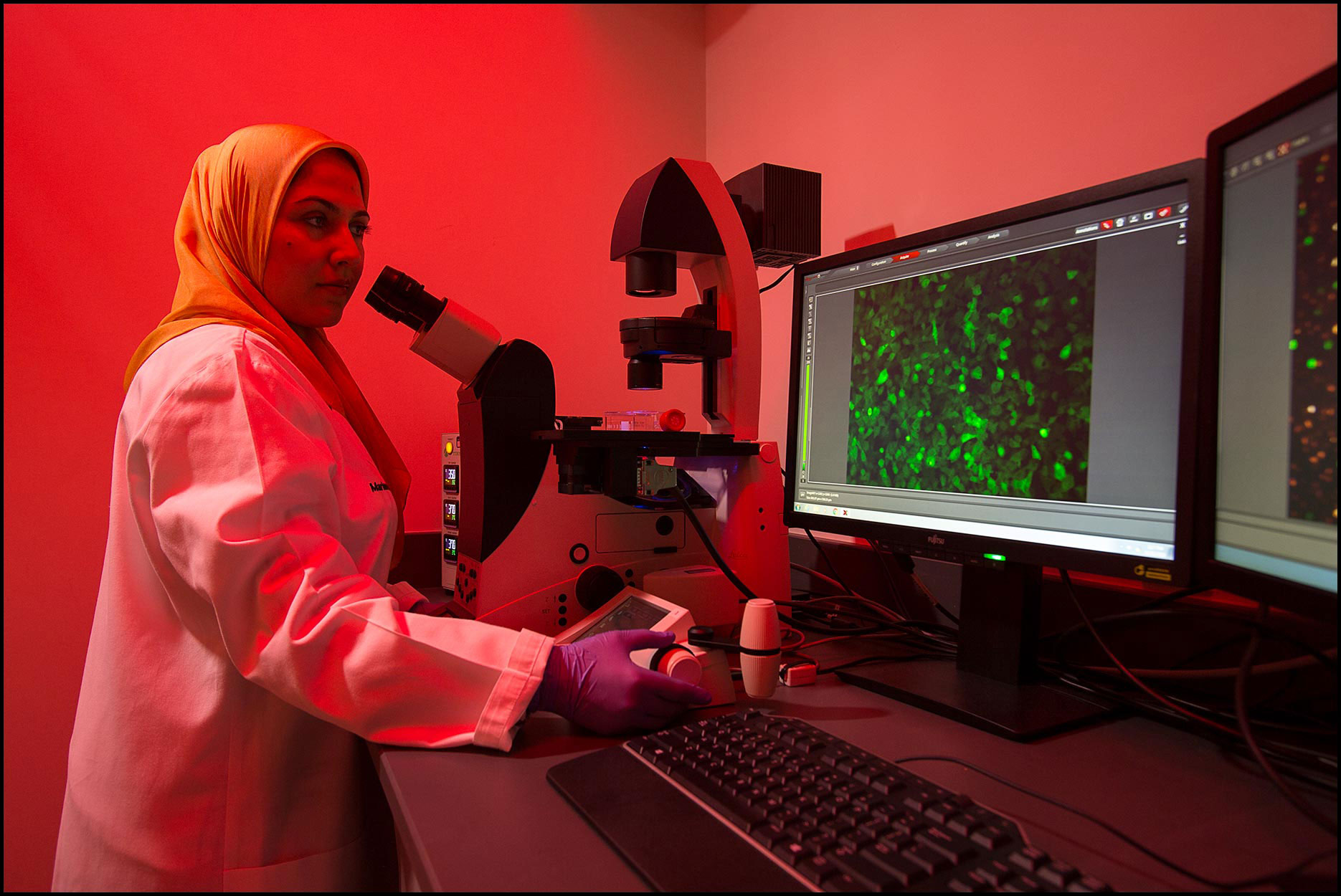A pharmaceutical researcher in a hijab and lab coat works in a microscope room with red ambient lighting.