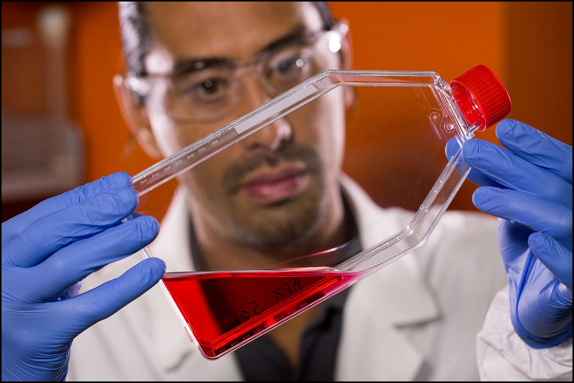 Detail of a lab technician in safety gear holding and examining a red liquid sample inside a plastic flask.