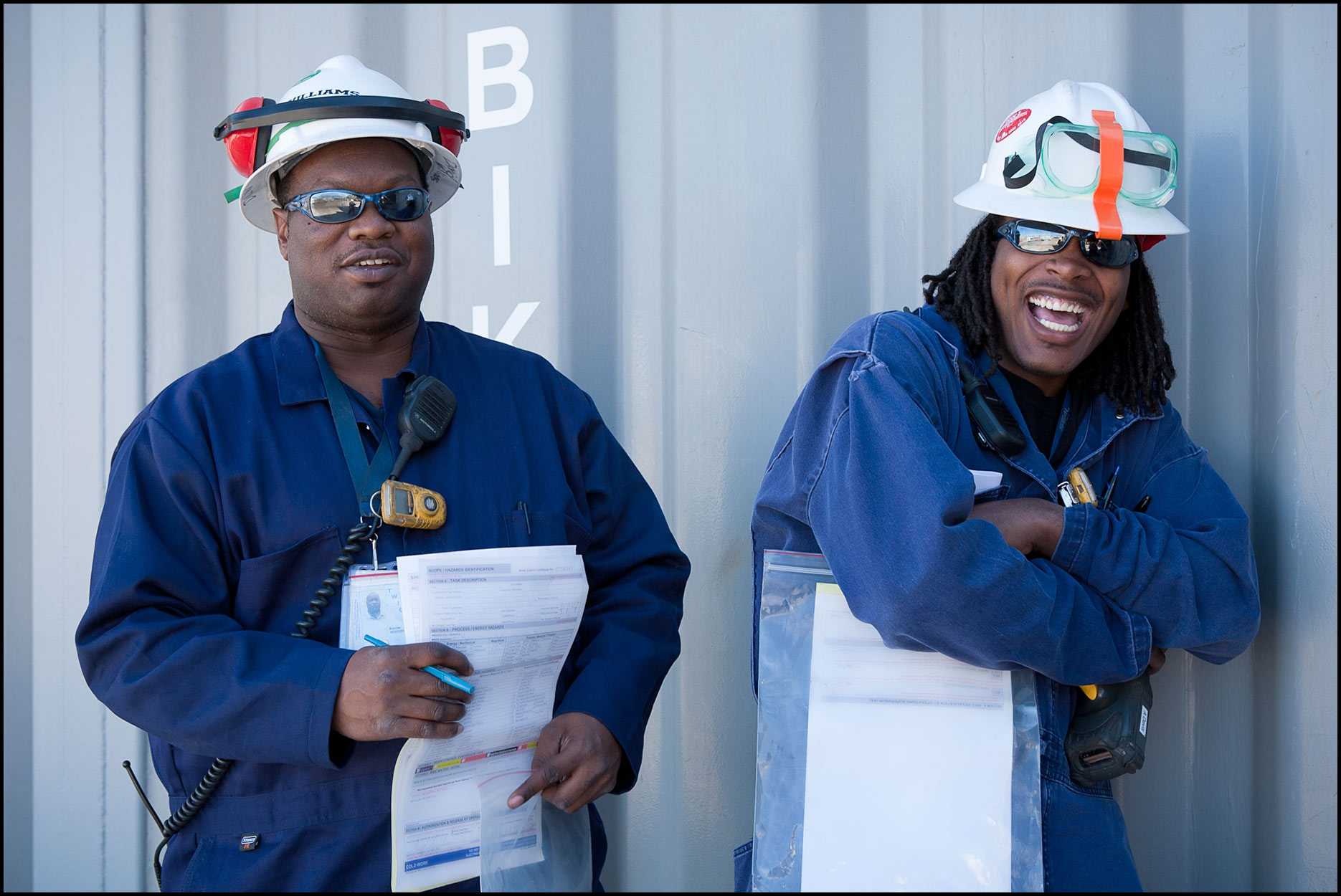 Two refinery workers in safety gear (PPE) have a laugh during a break from paperwork.