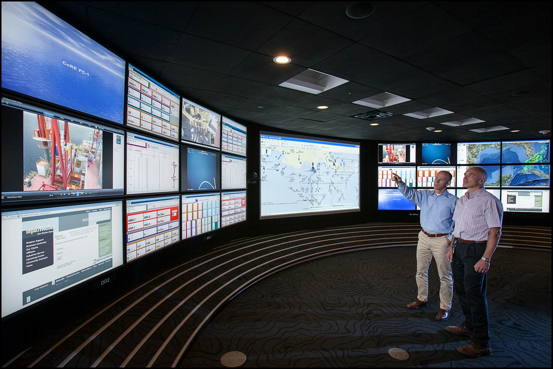 Two men watch an array of video displays monitoring offshore well activity.