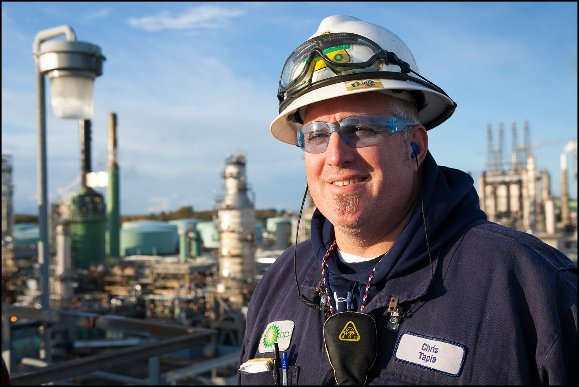 A portrait of a refinery operator wearing blue safety glasses and standing on top of a refinery unit.