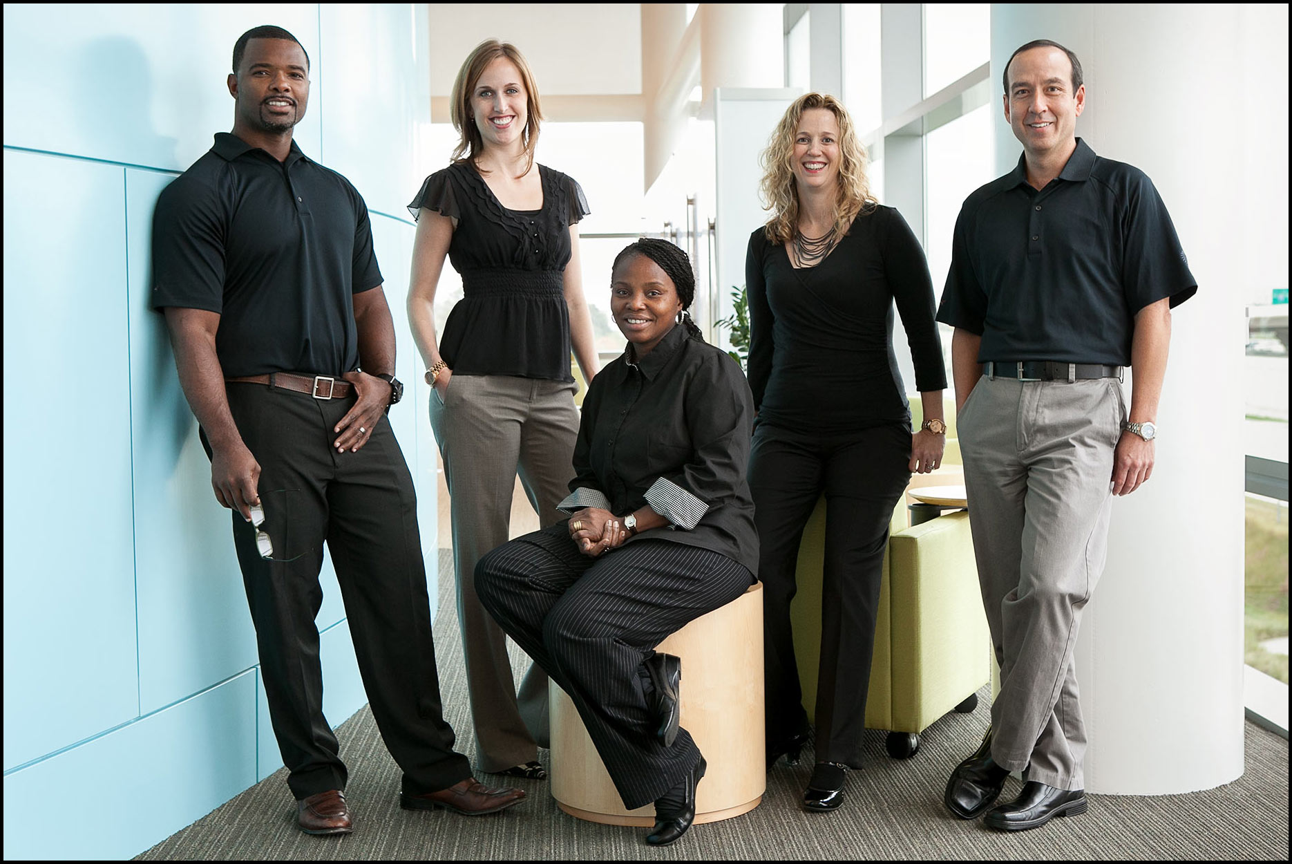 Management team, in business casual dress, stands in an office atrium for a group photo.