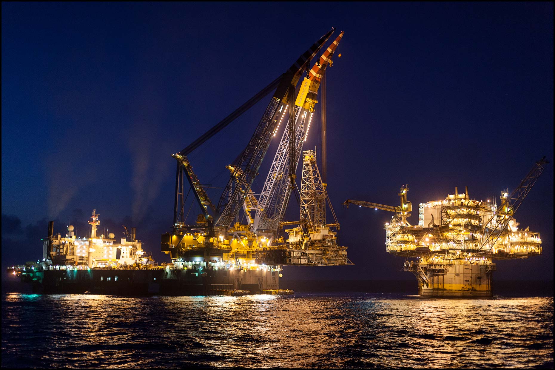 A construction ship lifts a drilling module onto spar rig structure at night.