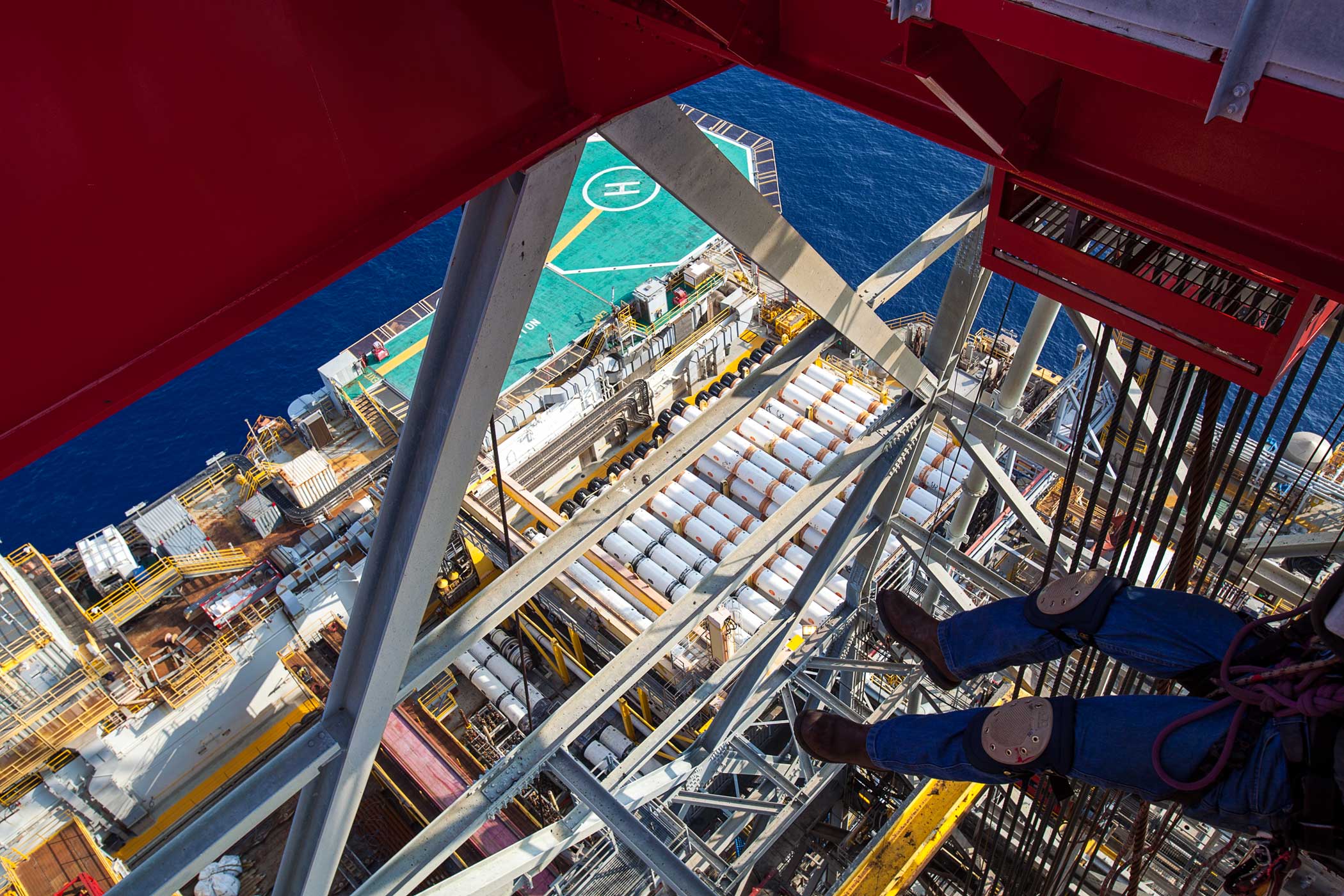 An overhead view of the legs and harness of an industrial painter suspended from the underside of an offshore rig derrick, high above the deck below.