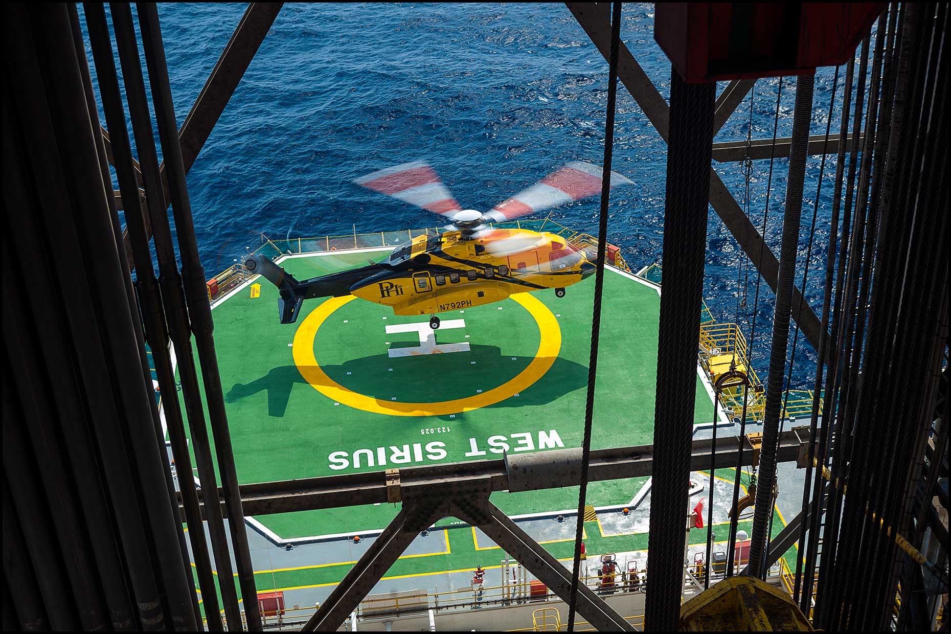 A crew helicopter lands on the helipad of an offshore drilling rig as seen through pipe stacks in the derrick.
