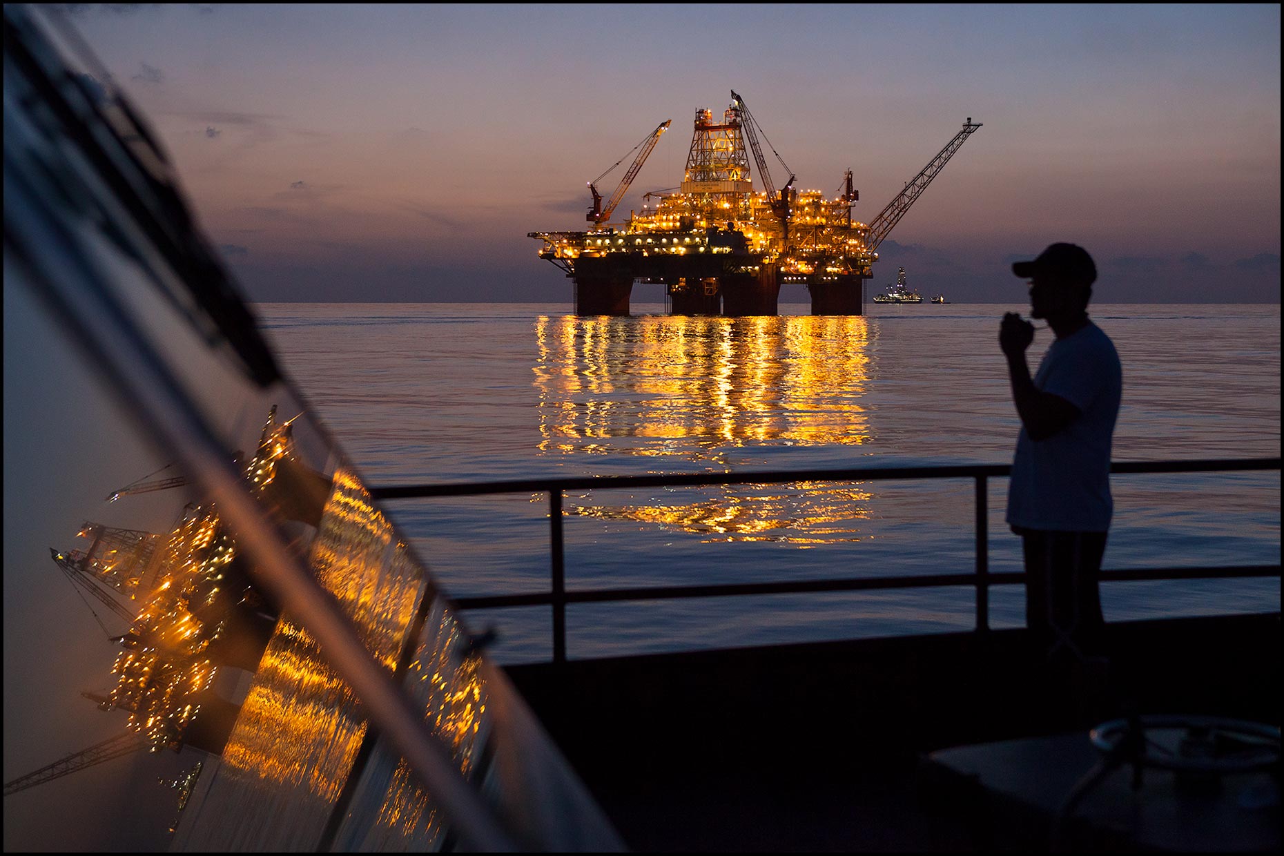Silhouette of the first mate standing on the deck of a supply vessel at twilight with a large offshore rig in the distance.