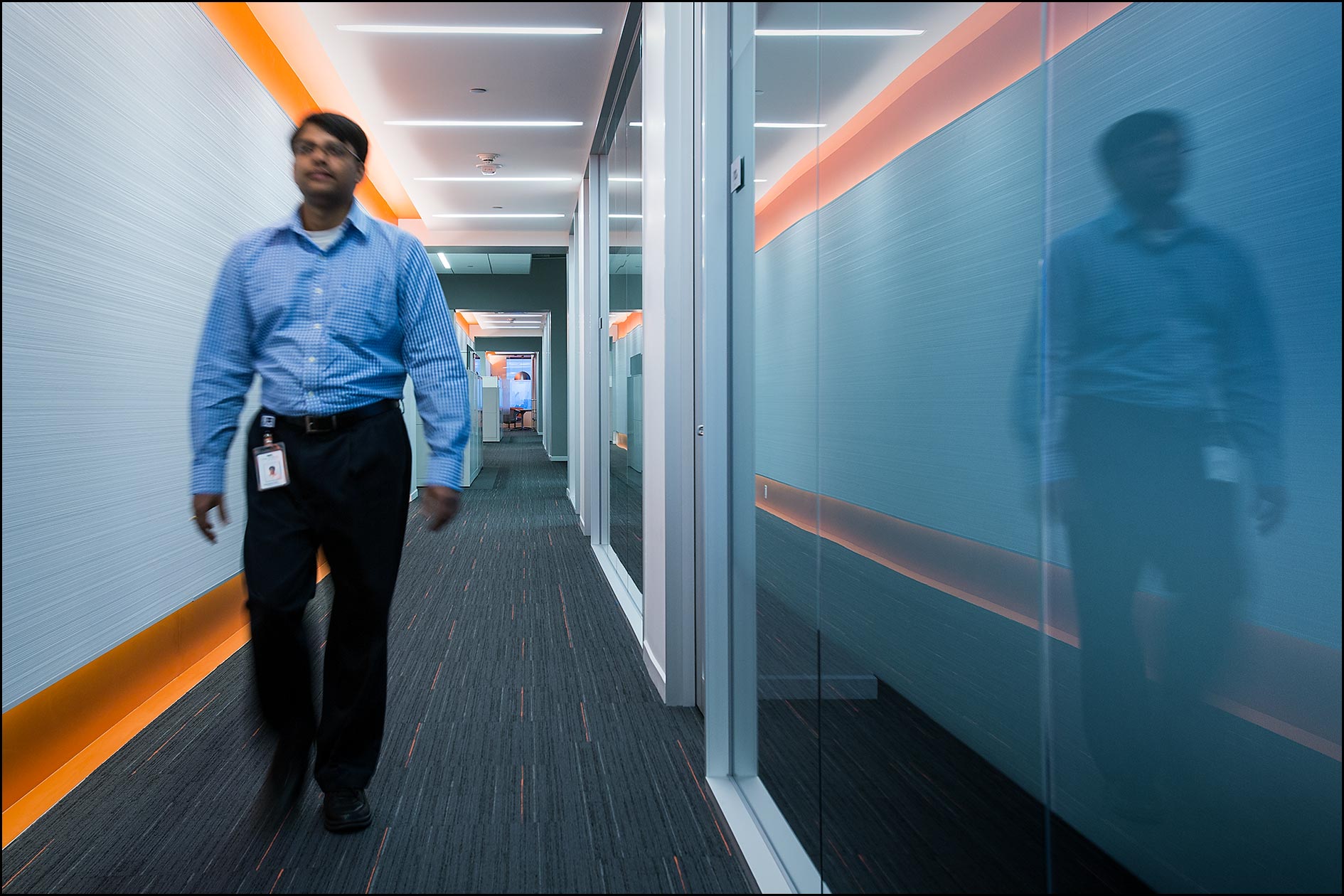 A male employee walks in a hallway with contemporary architectural decor with his reflection in a glass window.