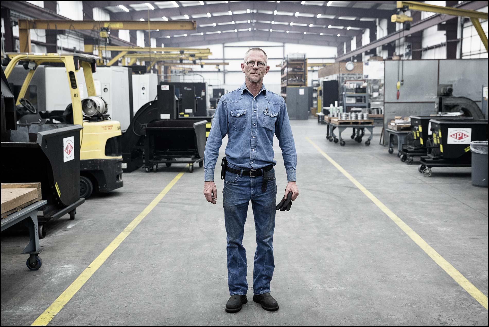 A portrait of a machinist standing in the center of a large machine shop.