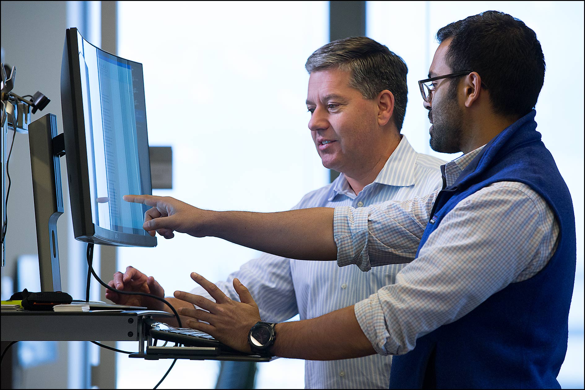Two employees analyze data at a computer on a standing desk.