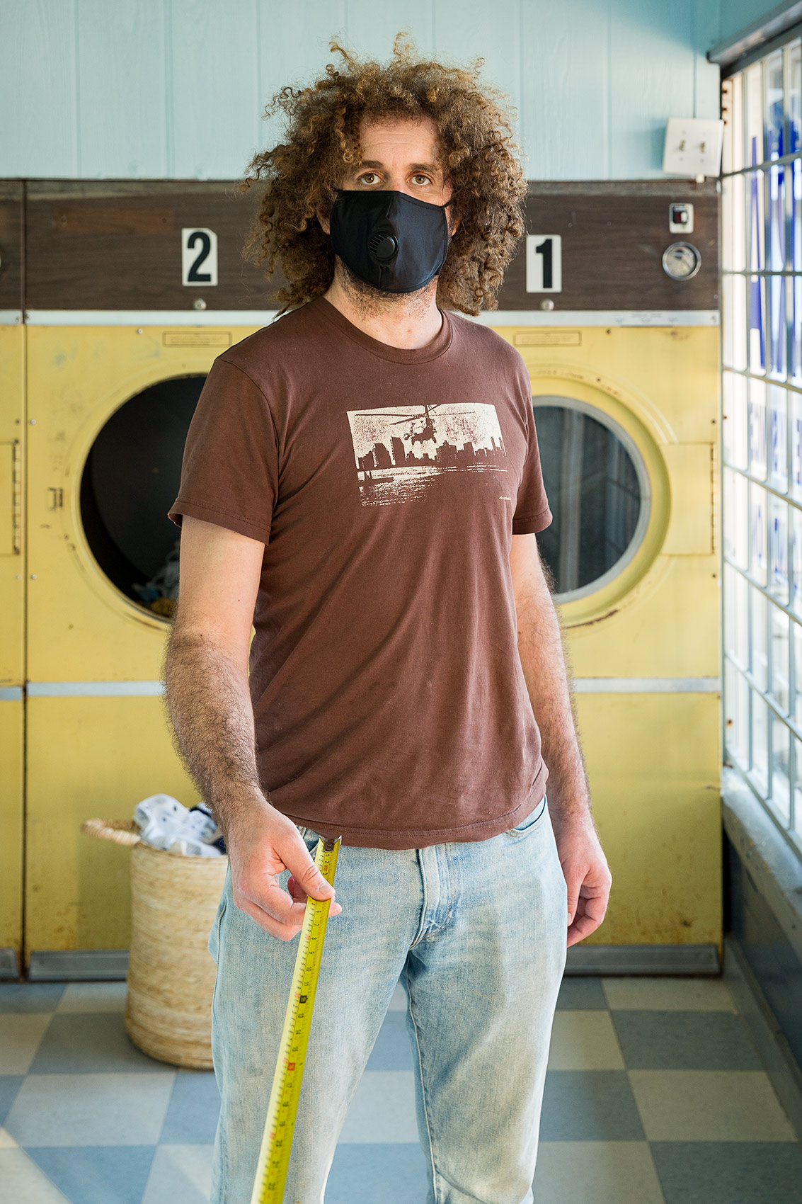  A man wearing a black mask stands inside a laundromat and holds a long tape measure that is a symbol of Covid-19 social distance