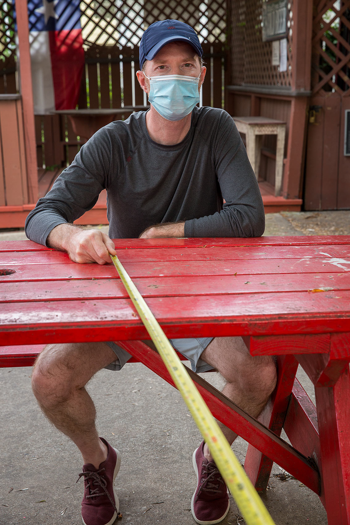 Man wearing a mask sits at a red picnic table with a long tape measure that is a symbol of Covid-19 social distance