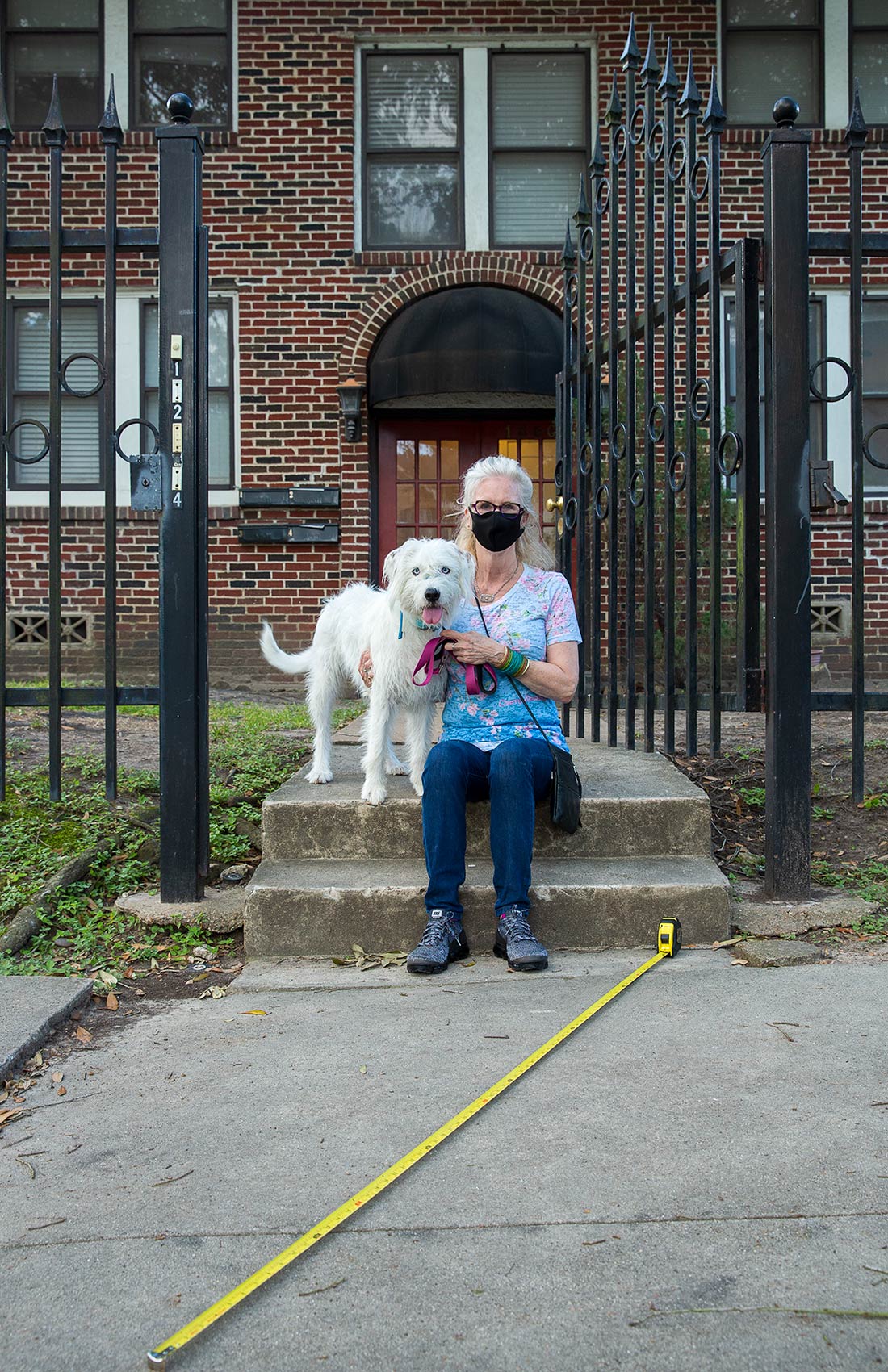 A masked woman and her dog sit on her porch with a long tape measure as a symbol of Covid-19 social distance