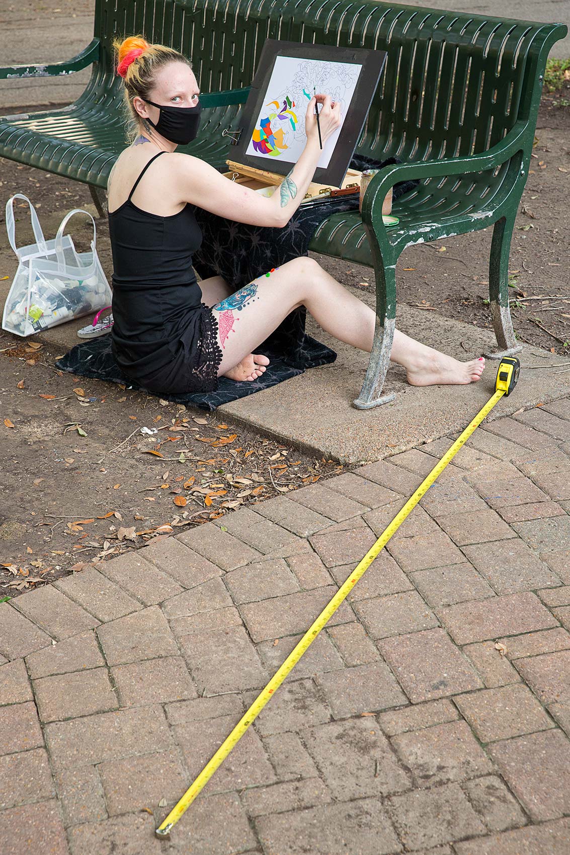 A young masked woman in a park paints on a canvas with a long tape measure as a symbol of Covid-19 social distance