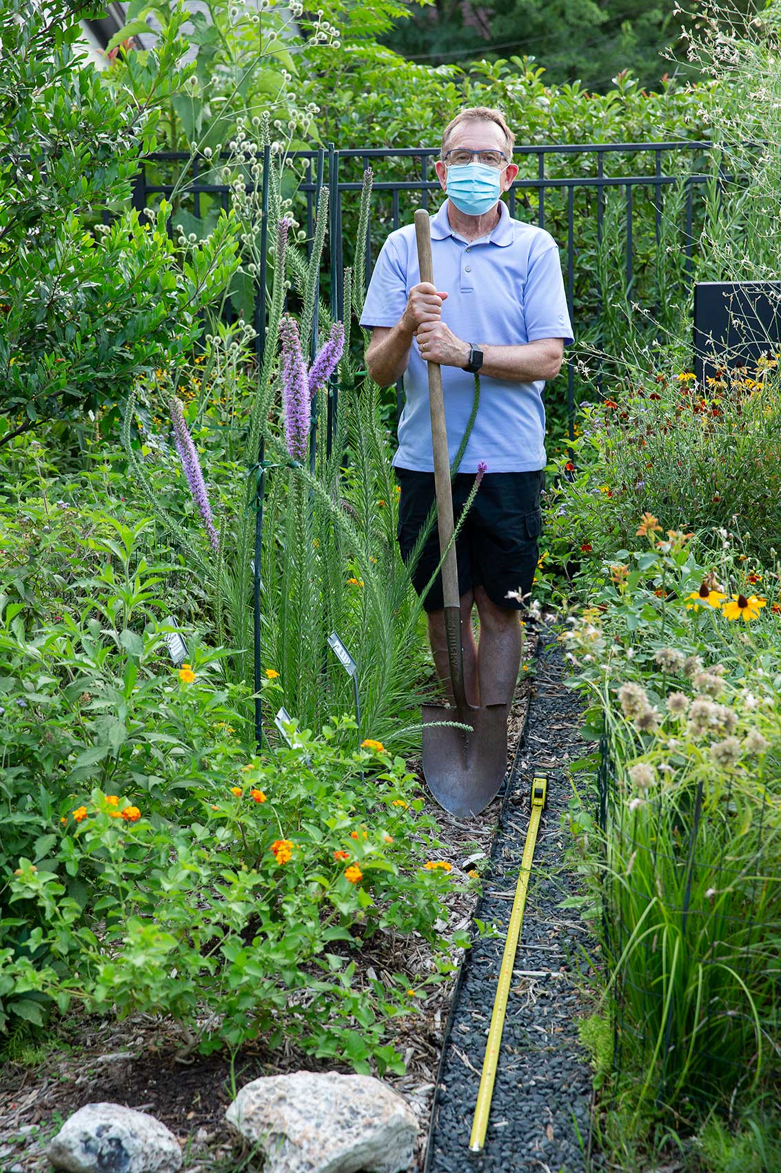 A man wearing a mask and holding a shovel works in his garden with a long tape measure as a symbol of Covid-19 social distance