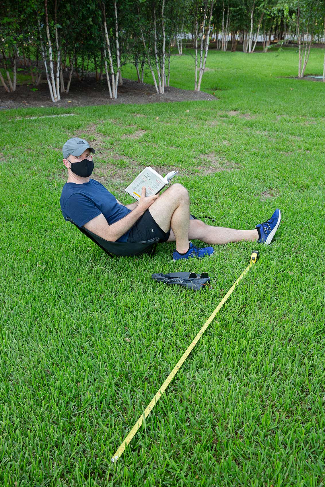 A man wearing a mask reads a book on a grassy lawn with a long tape measure as a symbol of Covic-19 social distance
