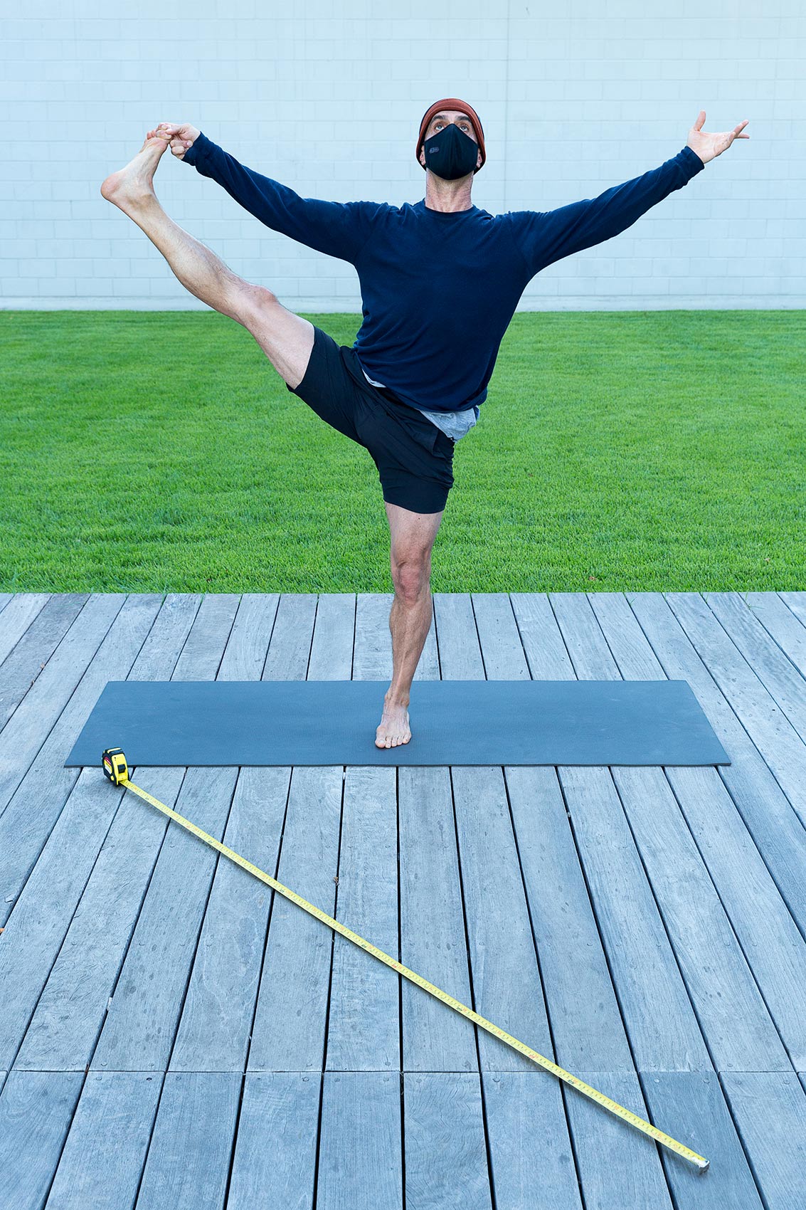 A man wearing a mask does a yoga pose on a wooden deck with a long tape measure as a symbol of Covid-19 social distance