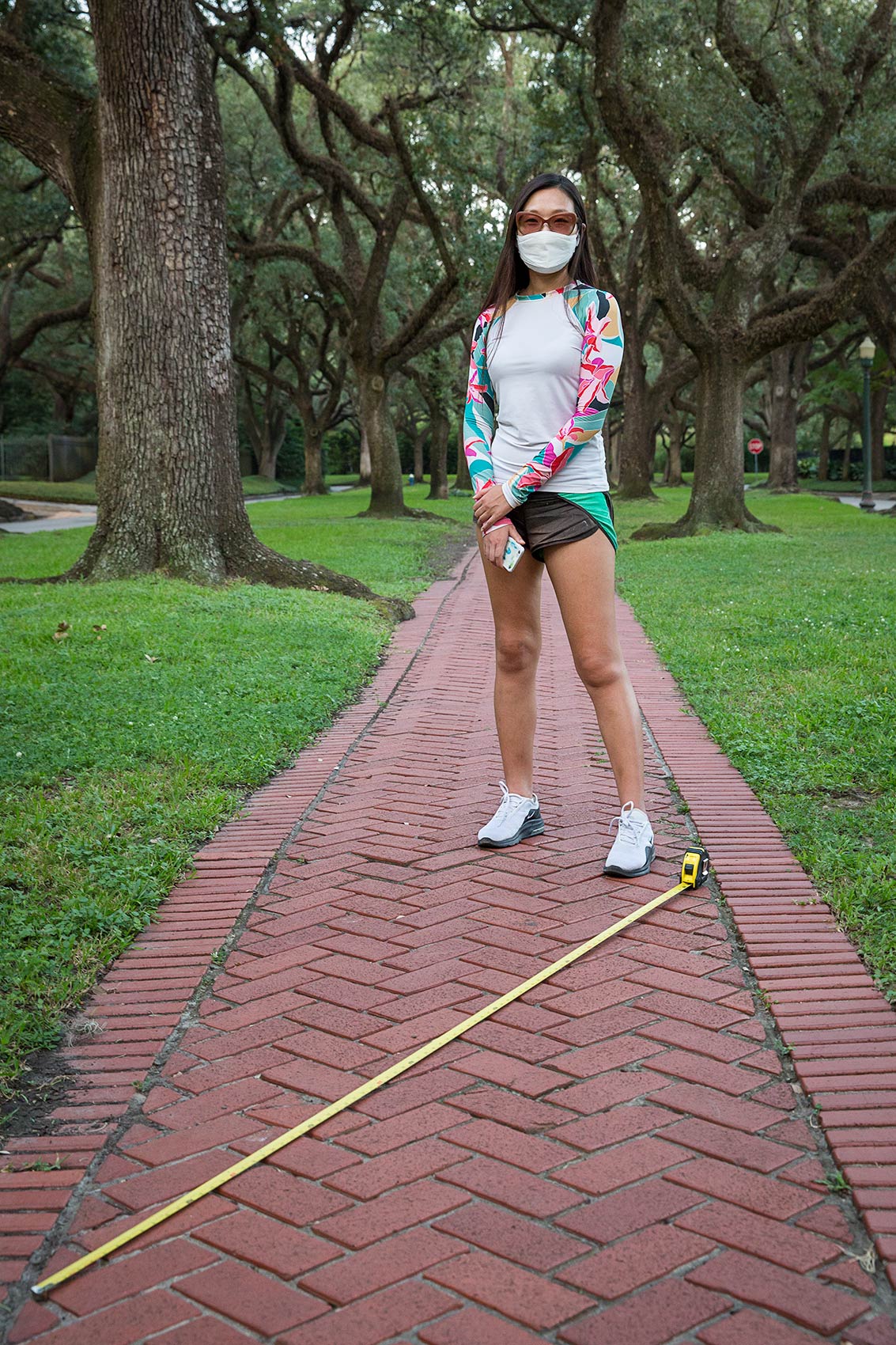 Young woman in mask on tree-lined walkway with long tape measure as a symbol of Covid-19 social distance
